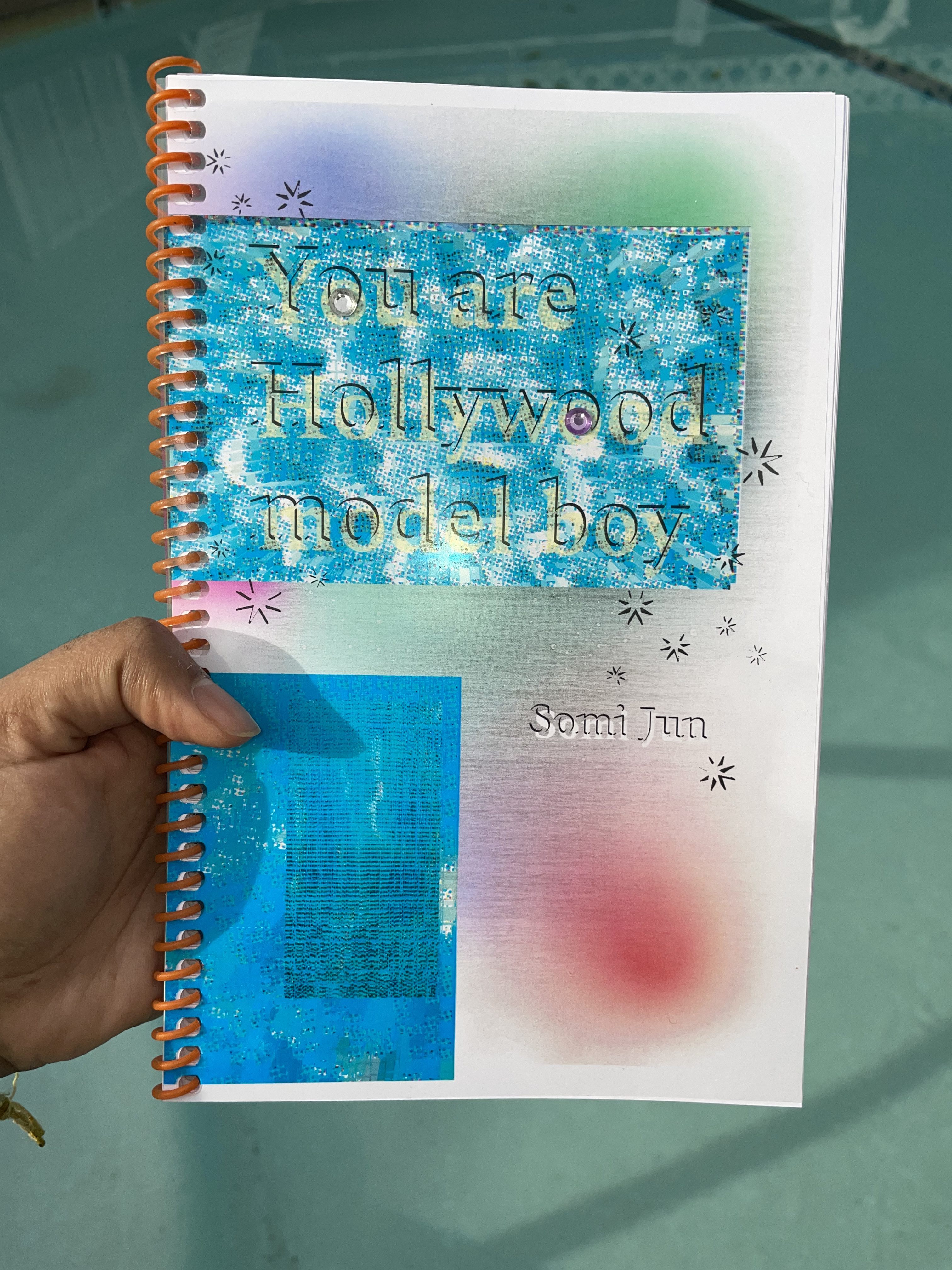 a spiral bound book with a blue, green, red, and purple cover that reads 'You are Hollywood model boy' is held out over a swimming pool